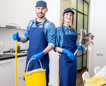 Portrait of a couple as a professional cleaners in uniform standing together with cleaning tools indoors Portrait of a couple as a professional cleaners in uniform standing together with cleaning tools indoors